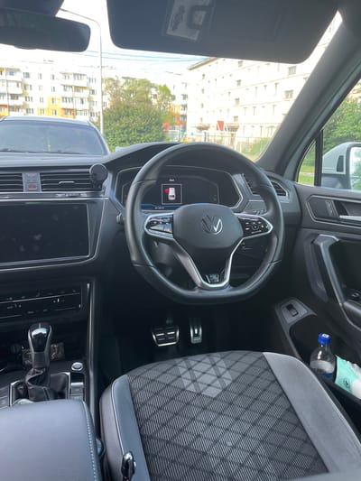 The image shows the interior of a Volkswagen Tiguan, likely in Sibirsky, Russia, based on the context provided. The car is parked, with the engine off, as indicated by the "ENGINE START STOP" button being illuminated. The interior features a black leather steering wheel with a Volkswagen logo, a digital dashboard displaying the time as 18:43 and trip information, and a central infotainment screen. The gear shifter is in the "P" (Park) position. The seats are upholstered in a dark fabric with a geometric pattern. On the passenger side, a water bottle and a packet of wet wipes are visible in the door pocket. Outside the car, apartment buildings and trees are visible, suggesting an urban or suburban setting. The lighting indicates it is late afternoon or early evening.