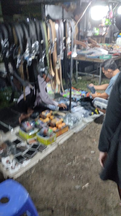 The image depicts an outdoor market stall, likely in Sukodono, Indonesia, at night. The scene is dimly lit, with bright artificial lights illuminating the area. The primary focus is on a stall selling a variety of goods, including a display of belts hanging on a rack and various packaged items laid out on a table and the ground.
Several people are visible. In the foreground, a person in a dark garment stands with their back to the camera. To the left, a person wearing a light-colored shirt and dark pants is crouched down, appearing to be interacting with the merchandise. To the right, another person in a blue shirt and patterned pants is seated or kneeling, also engaged with items on the stall. The atmosphere appears to be that of a busy market, with individuals browsing and making purchases.
The merchandise includes an assortment of belts with various buckle designs, neatly arranged. Other items are displayed in boxes and on trays, some appearing to be small electronic accessories or trinkets, judging by the packaging. The ground is covered with dirt and scattered debris, typical of an outdoor market setting. The overall impression is one of a vibrant night market scene. No visible text is discernable in the image.