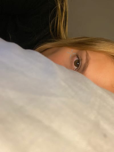 The image shows a close-up of a person's face, partially obscured by a white cloth. The visible portion of the face includes one brown eye with a cross drawn above it in a blue color. The person has blonde hair and fair skin. The background is blurred, with dark shadows on the right and a light gray wall visible in the distance. There is no evidence to suggest the location of Boulogne Sur Mer, Argentina.