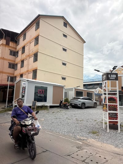 A person rides a motorcycle on a paved road in front of a building complex. The person is wearing a purple shirt and black shorts, and is riding a black motorcycle with a basket in the front. The building complex has several floors and is painted in light yellow and orange colors. In front of the building, there are several small, modern modular buildings, including a barber shop with a red and white striped pole. A silver electric car is parked nearby. A signpost with the text "S-BOX PROJECT" is visible. The ground is covered with gravel. The sky is cloudy. This scene is in Nonthaburi City Municipality, Thailand.