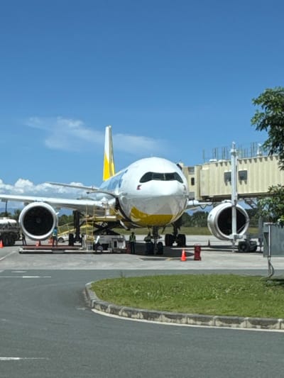 The scene depicts a wide-body passenger aircraft, featuring a white fuselage, yellow belly, and yellow tail fin consistent with Cebu Pacific livery, parked on an asphalt tarmac at Bohol-Panglao International Airport (TAG) in Panglao, Philippines. The aircraft's nose is oriented towards the viewer. A beige jet bridge is connected to the aircraft's forward left door.

Ground support equipment and personnel are present around the aircraft. A white fuel tanker truck, bearing "ARON" text, is positioned near the left wing. A white and gray cargo loader, with its platform elevated, is situated beneath the fuselage, between the left engine and the nose landing gear. At least two ground crew members are visible near the left engine, one in a light blue vest. Two additional crew members, wearing high-visibility yellow-green vests, stand near the nose landing gear. An orange traffic cone is placed on the tarmac in front of the right engine, with another cone and a red ground unit located further to the left.

The sky is clear blue with minimal scattered white clouds. The foreground consists of a curved asphalt road separated from the tarmac by a grassy median. Distant green trees and hills are visible in the background under the horizon line.