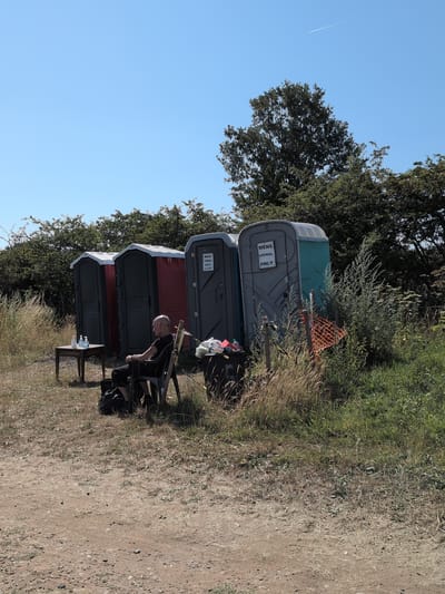 A man sits in a black chair outside several portable toilets under a clear blue sky in Claverley, United Kingdom. He is bald and wearing a black t-shirt and pants. In front of him is a small wooden table with two bottles of hand sanitizer. To his right, there is a large black trash can with some items in it. Behind him, there are four portable toilets. The first is grey, the second is red, the third is grey and labeled "MENS URINAL ONLY PLEASE", and the fourth is teal and grey. There is tall, dry grass and green bushes surrounding the area, with trees in the background. The ground is dirt and gravel.