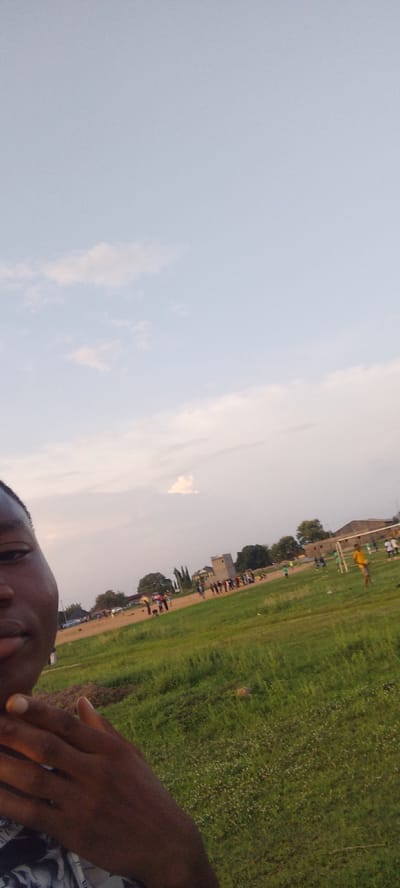 The image presents a selfie of a dark-skinned person, with part of their face and hand visible in the lower left corner. The background shows a grassy field with people gathered, some appearing to play soccer, indicated by the presence of a goal post. A building is visible further back, in the background. The sky is partially cloudy. The location appears to be in Ibo, Nigeria, inferred from the query context.