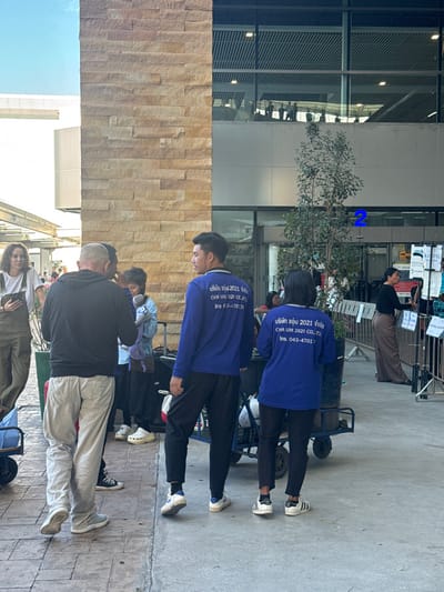 A group of people stand outside a building. Two men in blue shirts with white text on the back are prominent. The text includes "บริษัท ซอิ่ม 2021 จํากัด CHA UM 021 CC" and "Ins 03-470:70". One man is wearing a black shirt and light-colored pants. The building has a modern architectural style with glass windows. There is also a small tree in the background.