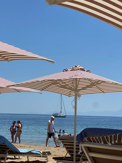 The image depicts a sunny day at a beach in Troulos, Greece. Several beach umbrellas with red and white stripes are set up on the sand. In the foreground, two lounge chairs are visible, one with a blue towel draped over it. The turquoise sea is calm and clear, with a few people swimming and a sailboat in the distance. Two women are standing in the shallow water, and a man is walking on the beach carrying a plastic bag. The sky is a clear blue, and mountains can be seen in the background.