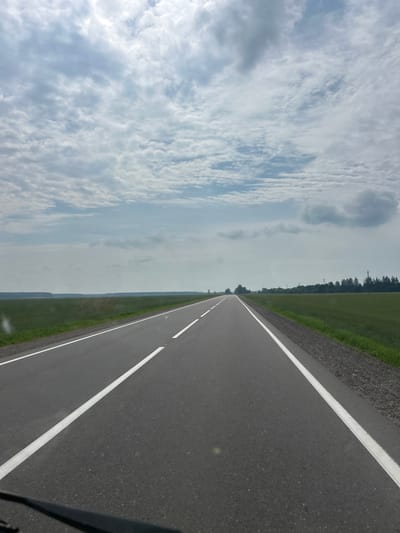 This media file shows a view from a vehicle traveling on a paved road in Дубненскі сельскі Савет, Belarus. The road has white dividing lines and a gravel shoulder. To the left of the road is a green field, and beyond that, a line of trees. The sky is filled with white, fluffy clouds, suggesting it is daytime and the weather is fair. The perspective suggests the photo was taken from the passenger seat of a car. There is no visible text in the image.