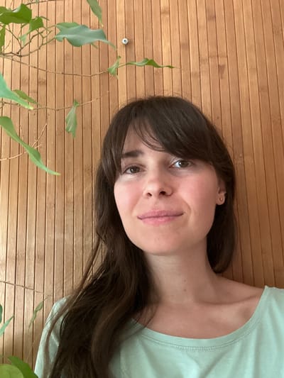 The image is a close-up, head-and-shoulders portrait of a woman with dark brown hair and bangs, looking directly at the camera. She has fair skin, light brown eyes, and is wearing a light green t-shirt. She has a small gold stud earring in her right ear. She is positioned in front of a wall constructed of vertical bamboo slats. To the left of the woman, a plant with green leaves and thin branches extends into the frame from the top left corner. There is a small silver screw visible on the bamboo wall behind her head. The image appears to be taken indoors. The specific location in the city of Chornomorske, Ukraine, is not determinable from the visual information provided in the image.