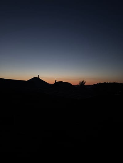 The image shows a silhouetted landscape at dusk. The foreground is dominated by the dark shapes of rooftops, casting long shadows. Beyond the rooftops, the sky transitions from a deep blue at the top to a pale orange and yellow near the horizon, indicating the setting or rising sun. A few faint stars are visible in the upper part of the sky. In the middle ground, silhouetted trees and a single tree with more detail can be seen against the lighter sky. The overall impression is one of quiet and stillness.