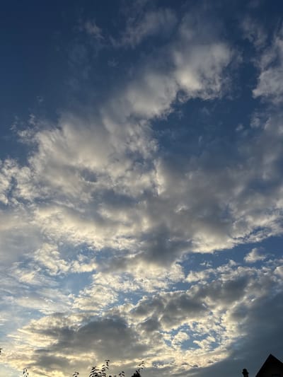 A low-angle shot captures a vast expanse of the sky over Мартемьяново, Russia, filled with dynamic clouds. The sky is a deep blue, punctuated by a variety of cloud formations, ranging from wispy, scattered white clouds to larger, more dense gray masses. Some clouds are illuminated by sunlight, appearing bright and almost glowing, while others are darker and cast shadows. The bottom of the frame shows the silhouettes of tree branches against the bright sky, and a corner of a dark-colored building, possibly a house with a chimney, is visible on the far right. The overall impression is of a dramatic and atmospheric skyscape.