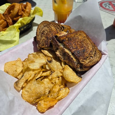 This is a close-up, eye-level shot of a meal served in a restaurant in Cannon Beach, United States of America. The meal consists of a grilled sandwich and a side of potato chips, presented in a white paper-lined basket with a pink paper liner. The sandwich, cut in half diagonally, is made with dark bread and appears to have a thick patty and melted cheese, with a generous amount of sauce oozing out. The potato chips are golden brown and appear to be kettle-cooked. In the background, out of focus, is a basket filled with golden-brown onion rings, a glass of orange-colored liquid with ice and a garnish, and a sliver of a red and white sign. The scene is lit by natural sunlight, casting shadows on the table surface.