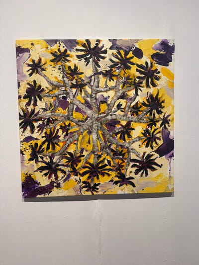 This is a square, abstract painting with a textured, impasto technique. The artwork is dominated by a vibrant yellow background, overlaid with strokes of purple and grey. The central subject appears to be a stylized representation of a plant or tree, with dark purple, star-shaped floral elements radiating outwards from a grey, branching structure. Splashes and drips of black paint are visible throughout the composition, adding to the abstract and energetic feel. The painting is hung on a white, textured wall, which is slightly uneven, suggesting it might be in a gallery or studio space. The lighting appears to be artificial, casting a slight shadow to the right of the artwork. There is no visible text within the image. The overall impression is one of bold color and dynamic form, possibly evoking a sense of nature viewed through an expressive and non-realistic lens.