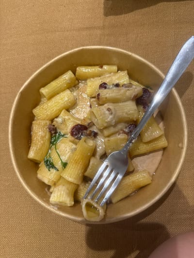 A close-up, overhead shot shows a bowl of rigatoni pasta with a creamy sauce, dark red berries, and green herbs. A silver fork rests in the pasta, with one prong inside a tube of pasta. The pasta is in a brown paper bowl on a textured, brown tablecloth. The scene is lit from above, casting a shadow of the bowl and fork onto the tablecloth. No specific location in Mont-Saint-Guibert, Belgium is discernible from the image.