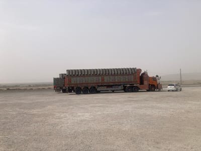A heavily loaded semi-truck, decorated with colorful patterns, sits in a barren landscape alongside a small white car. The truck is full of cylindrical cargo. The scene is washed in hazy light, suggesting either dust or fog. A utility pole stands in the background. The ground is light-colored and appears arid.