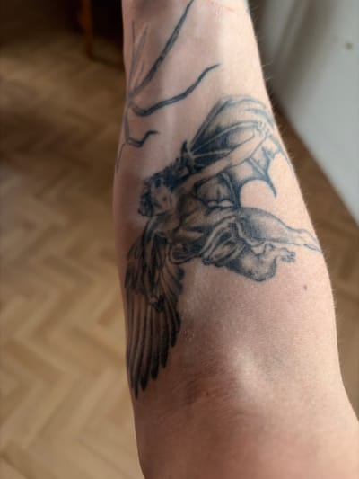 The image shows a close-up of a tattoo on a person's arm. The tattoo appears to be an artistic depiction of a winged figure, possibly an angel or a demon, in a dynamic pose. The style is black and grey, with intricate shading and linework. The background is blurred, showing what looks like a wooden floor and a wall, suggesting an indoor environment. There is no visible indication of the location being in Göttingen, Germany. The subject is a human arm with a detailed tattoo.