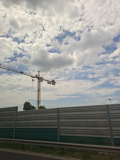 A construction crane with a yellow lattice tower and a long horizontal boom is visible against a cloudy sky. The crane has a cabin for the operator and a hook suspended from the boom. In the foreground, a metallic sound barrier with horizontal slats runs across the frame. The barrier is divided into two sections: the upper part is silver, and the lower part is dark green. A grey metal guardrail is positioned in front of the sound barrier, with a strip of grass and dirt beyond it. In the background, trees can be seen behind the sound barrier, and a lamppost is visible on the right side of the frame. The scene appears to be located on the outskirts of a city, possibly Dybów Kolonia, Poland, given the presence of construction and infrastructure elements. There are no people or animals visible in the image. The lighting suggests it is daytime, with overcast conditions.