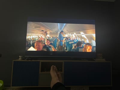 The image shows a television screen displaying a scene from inside an airplane. The airplane cabin appears crowded with passengers seated and a flight attendant in a blue uniform standing in the aisle. The lighting suggests it is either dawn or dusk, with warm light filtering through the airplane windows. Below the television, a dark blue and light gray TV stand is visible, with a person's bare feet resting on the floor in front of it. The environment is dimly lit, indicating an indoor setting, likely a living room in Kaipy, Russia.