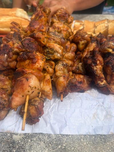 A close-up, slightly elevated view shows several skewers of grilled chicken, seasoned with herbs, arranged on a white paper covering. In the background, there are sliced portions of bread. A person's hands are visible in the upper portion of the image, appearing to be in the process of handling or serving the food. The scene appears to be outdoors, possibly at a picnic or event, with a hint of green foliage in the background. The chicken pieces are varied in size and color, with some showing charring from the grill. The bread appears to be a rustic loaf. The overall impression is of a casual outdoor meal being prepared or served. The location is identified as Campezo/Kanpezu, Spain.