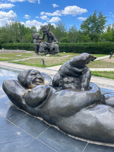 The image depicts a bronze sculpture of a woman lying down while holding a child above her. The sculpture is located in Changping District, China, in a park-like setting with green grass and trees in the background. Other sculptures of people sitting are visible in the distance. The sky is blue with white clouds. The sculpture is situated on a tiled surface that reflects the sky.