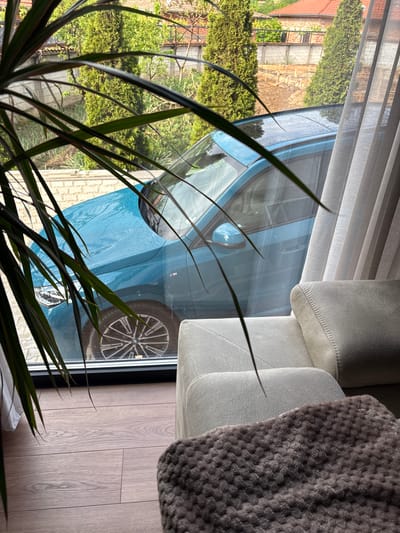 The image shows an interior view with a focus on a window reflection. A blue car is visible through the window, reflected among the interior elements. A white couch with a gray throw blanket is partially visible on the right side of the frame. Green plants are present in the lower-left corner. The view outside the window reveals parts of a building, trees, and possibly a brick walkway. The location is Dolno Drianovo, Bulgaria.