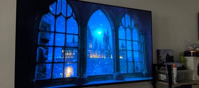 This is a horizontal, eye-level, close-up shot of a television screen displaying a scene from the movie Harry Potter and the Prisoner of Azkaban. The screen shows a view through a gothic-style window frame, with two arched panes looking out onto a snow-covered castle. The castle is illuminated by a bright, full moon and scattered lights from within its many windows. The scene is depicted in a dark blue hue, with white specks representing falling snow. In the foreground, on the left side of the screen, a lit candle in a holder is visible through the window pane. The television itself is dark and positioned on a surface that appears to be a TV stand. To the right of the television, on the wall, is a framed picture and some objects on a shelf, including a green toy dinosaur and a white aerosol can. The background on the left appears to be a textured wall. The scene is set at night. There is no visible indication of the location being Villeneuve-le-Roi, France.