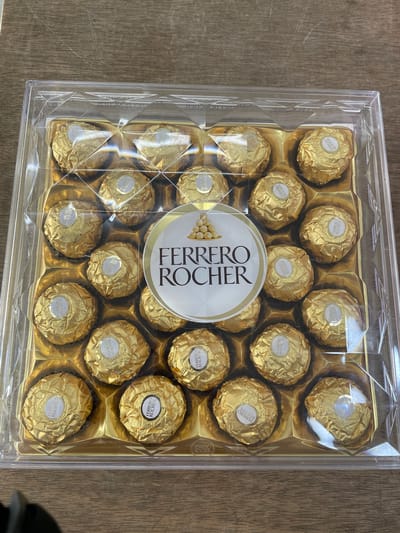 An overhead shot captures a clear plastic box containing an arrangement of Ferrero Rocher chocolates. The chocolates, each individually wrapped in golden foil, are neatly placed in a grid pattern within the box. A prominent circular label with the "Ferrero Rocher" logo is centered within the chocolates, adding a brand identifier. The box rests on a wooden surface. The location of San Jose del Monte, Philippines, is not discernible from the visual information provided in the image.