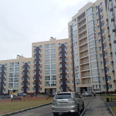 The image shows a residential complex in Arseniev, Russia. The complex consists of several multi-story buildings with beige and dark brown facades, and numerous windows and balconies. In the foreground, a silver SUV is parked on a paved road. In the background, a playground with swings and slides is visible. A few people are seen walking on the paved pathway. The sky is overcast, suggesting a cloudy day.