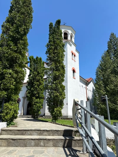 The image shows a white church with red brick accents in the city of Barziya, Bulgaria. The church is partially obscured by tall green trees. A set of stone steps with a metal railing leads up to the church. The sky is clear and blue.