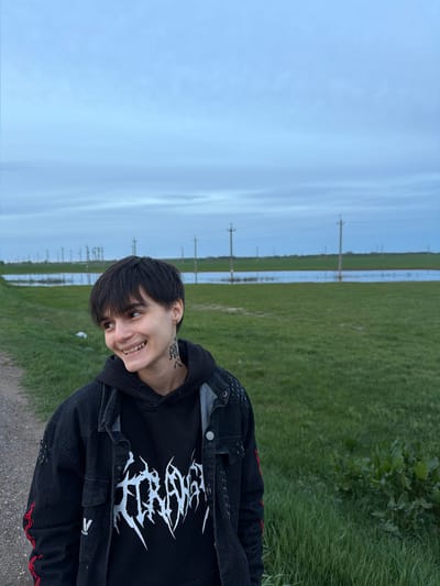 A photo taken in Azyakul', Russia, depicts a person standing in a grassy area. The sky is overcast. Power lines are visible in the background. The person is wearing a black hoodie with a white graphic, and a black denim jacket with red stitching. They have short, dark hair and a tattoo on their neck. They are smiling.