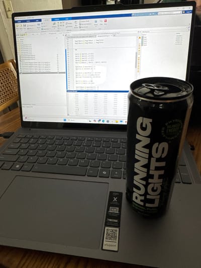 A can of "Running Lights" energy drink rests on a gray Lenovo Yoga laptop. The laptop's keyboard is visible along with a sticker featuring the word "Yoga", a screen size of "13.3in" and various icons with text such as "2K CAFFEINE FREE". The can is black with the brand name in bold white letters. The laptop screen displays an interface with code, likely MATLAB. The overall scene suggests a workspace or desk setting.