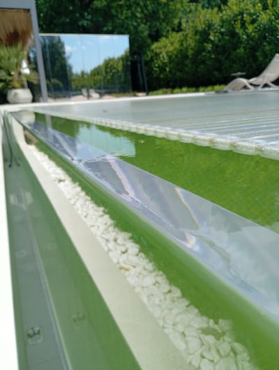 The image depicts a close-up, angled view of an outdoor swimming pool in Nowa Wieś Szlachecka, Poland. The pool has a modern design with clean lines and a white outer edge. Inside the pool, the water is a vibrant green color, suggesting it may contain algae or a special treatment. A clear, reflective cover lies partially over the pool, creating a corrugated surface. Beside the pool, small white pebbles are visible in a decorative channel. In the background, lush green trees and a clear blue sky are reflected in a large, sleek glass panel. A portion of a light-colored lounge chair is also visible in the reflection, hinting at a relaxation area. The overall impression is one of a well-maintained, contemporary outdoor space.