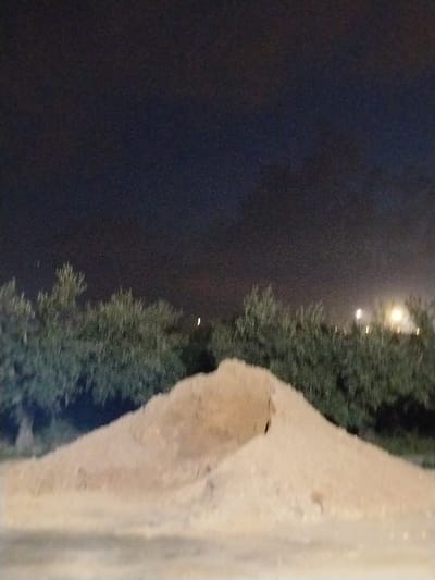 A low-lit landscape displays a sandpile as the primary focus. Behind the sandpile, a line of trees extends horizontally across the frame. Above the trees, the sky is dark, with subtle gradients of color suggesting it is night. Distant blurred lights punctuate the sky near the horizon, adding a sense of depth to the scene. The lighting emphasizes textures within the sandpile and foliage.