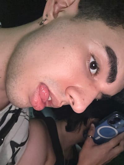 A close-up shot reveals a young man with dark hair and eyebrows, brown eyes, and fair skin. His tongue is extended, slightly protruding from his lips. He has multiple piercings on his ear. A blurred figure behind him holds a blue cell phone, partially obscuring the background. The man wears a white and black striped shirt. The image showcases the man's facial features and expression in detail.