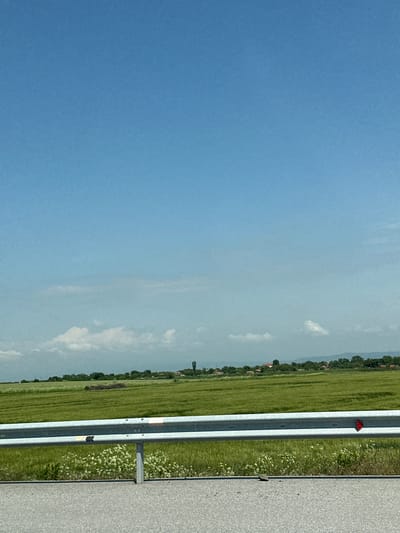 The image, taken in Malenovo, Bulgaria, depicts a sunny, clear day over a rural landscape. The dominant feature is a vast expanse of bright blue sky, with a few wispy white clouds scattered across it, particularly towards the horizon.
Below the sky, a wide stretch of lush green fields, possibly agricultural, extends into the distance. A line of trees marks the far horizon, interspersed with what appear to be small settlements or houses characterized by red roofs.
In the mid-ground, a metallic guardrail runs horizontally across the frame, dividing the scene. To the right of the guardrail, a section of grey asphalt road is visible. To the left of the guardrail, there is a grassy verge dotted with small white wildflowers, separating the road structure from the fields. A small red triangular reflector is attached to the guardrail in the upper right portion, and a grey support post is visible further down.
The time of day is bright daylight, indicating mid-day, with excellent visibility. No people or specific activities are present, portraying a calm, static landscape view, likely from alongside a highway or main road. No text is visible in the image.
