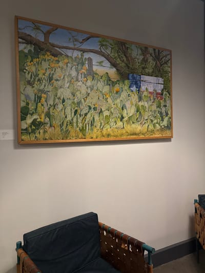 A framed painting depicting a Texas landscape hangs on a white wall. The artwork features a weathered building with a Texas flag visible behind a dense collection of prickly pear cactus. To the right of the painting, a section of modern furniture with leather and dark blue cushion accents is partially visible.