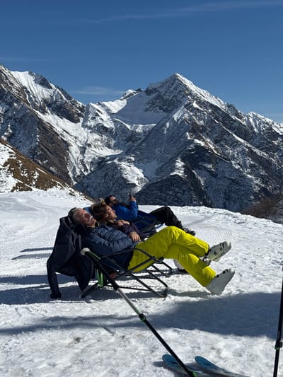 The image displays a group of people riding a ski lift in a majestic, snow-covered mountain range in Alagna Valsesia, Italy. The setting is a clear, sunny day, with a brilliant blue sky dominating the upper portion of the frame.
Five individuals are seated on a chairlift, ascending or traversing a ski slope. The person at the far end is wearing distinctive bright yellow ski pants and white ski boots, with the brand "HEAD" visible on one boot. Their skis and ski poles are seen standing vertically in the groomed snow to the side of the lift. The other passengers are dressed in various winter jackets, predominantly dark blue and black. One person in a blue jacket is actively pointing towards the panoramic mountain view, suggesting engagement with the stunning scenery.
The background features towering, rugged peaks covered extensively in snow, interspersed with rocky outcrops. The sunlight casts shadows, highlighting the contours and textures of the mountains. The ground below the chairlift shows a wide expanse of groomed snow, indicated by parallel tracks, confirming the location is an active ski resort. The overall atmosphere conveys a sense of serene enjoyment amidst the grandeur of the alpine environment.
