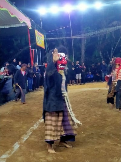 An outdoor night scene in Tamansari, Indonesia, depicts a cultural performance. The central figure, barefoot, wears a highly stylized red mask featuring large white eyes, white fangs, and black hair. This individual is dressed in a dark, long-sleeved top over a lighter garment and plaid-patterned sarong-style trousers. Their right hand is raised, fingers spread, while a long, light-colored object hangs from their left hand. To the right, a second barefoot performer, partially visible and blurred, wears a red head covering and striped clothing. The performance occurs on a dirt ground marked with a white line. Numerous spectators, casually dressed, stand in the background, observing the event. On the far left, a barefoot man in dark attire walks away from the center, carrying a stick. The area is lit by multiple overhead artificial lights. A pink tent canopy and a yellow banner with text are visible in the upper-left background, with trees in the distant background.