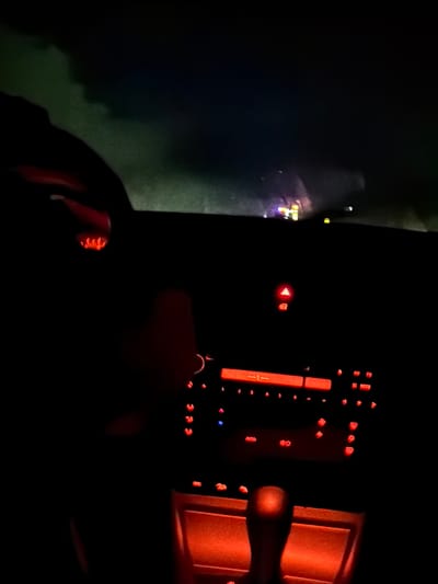 The image depicts the interior of a car at night, with the dashboard and center console illuminated by red ambient lighting. The focus is on the car's stereo system and controls, which are glowing with red light. The car appears to be in motion, as indicated by the blurred background showing distant lights and a dark, possibly overcast sky. The scene is set within the city of Tomai, Moldova, suggesting the vehicle is on a road within that location. There are no people clearly visible in the image, though a hand may be present on the steering wheel, partially obscured by darkness. The overall impression is one of a nighttime drive through an urban or suburban area in Tomai.
