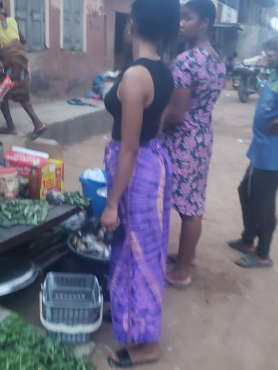 The image depicts a scene in what appears to be Alalubosa, Nigeria. Three people are visible. One woman, wearing a black top and purple patterned skirt, is handling items on a table or stand. On the table are food items, including leafy greens and boxed goods. Another woman, wearing a floral patterned dress, stands behind her. A man wearing a blue shirt and black pants is partially visible in the background, along with other individuals in the background. The scene appears to be outdoors, with buildings visible in the distance.