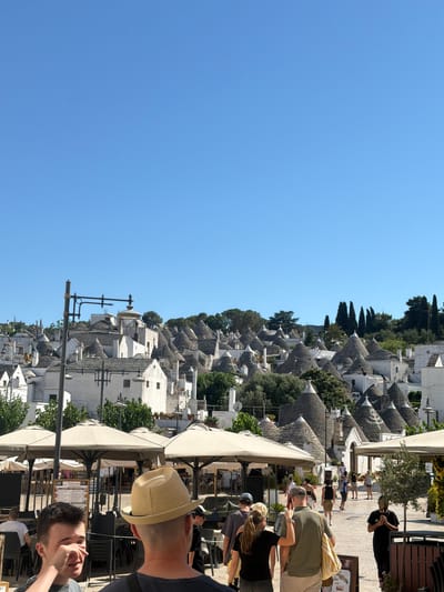 The image depicts a sunny day in Alberobello, Italy, a city known for its unique Trulli houses. In the foreground, several people are walking through an outdoor area with many tables and umbrellas, suggesting a marketplace or dining area. The iconic Trulli, with their conical roofs, dominate the background, creating a picturesque and historic cityscape. The sky is a clear, bright blue, and the overall atmosphere appears lively and tourist-friendly.