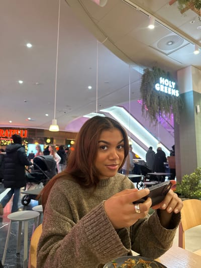 The image depicts an indoor scene within the Westfield Mall of Scandinavia, Solna, Sweden, specifically in a dining area.

In the foreground, a young adult female with medium brown skin and shoulder-length dark brown hair, parted centrally, is seated. She is wearing a loose-fitting, heathered olive-green knit sweater. Her gaze is directed towards the camera, and she exhibits a slight smile. She holds a dark-colored smartphone horizontally with both hands, her thumbs positioned over the screen. A prominent silver-toned ring with a textured surface is visible on her left ring finger. Her fingernails are manicured with a light-colored polish. A plate containing what appears to be a salad or shredded vegetable dish is partially visible on a light-colored table in the lower right.

The background reveals a bustling commercial environment. To the left, part of a red sign bearing the word "FRATELLI" is visible, indicating a restaurant. Several indistinct figures are walking in the mid-ground, some wearing dark winter coats, and a black baby stroller is present. To the right, a white neon sign displays "HOLY GREENS," positioned above a section of decorative hanging greenery. Behind this sign, an escalator with illuminated purple/pink undersides ascends to an upper level. Additional people are visible near the Holy Greens establishment and the escalator. The space is brightly illuminated by numerous recessed ceiling lights and some pendant lights, featuring a light-colored ceiling and modern architectural elements.