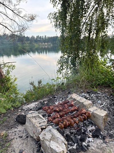 The image depicts an outdoor barbecue scene by a body of water, likely a lake or river, in Sulejów, Poland. In the foreground, skewers of meat and mushrooms are grilling over hot coals, supported by cinder blocks. The ground is covered with ash and debris from the fire. Behind the barbecue, lush green foliage and trees are visible, with a tranquil body of water reflecting the sky. The background shows a forested bank with a sandy cliff face. The lighting suggests it is either early morning or late evening. No people are present in the image.