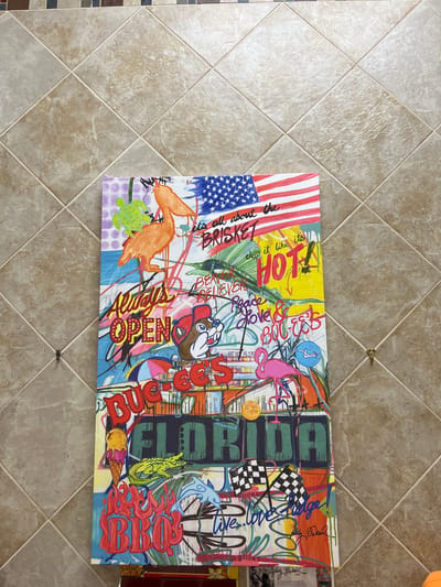 The image shows a colorful, vibrant painting leaning against a tiled wall. The painting is vertical and appears to be a mixed-media piece with elements of drawing and painting.
The painting's content is a collage of various images and text. At the top, there is a partial American flag. Below the flag, text reads "it's all about the BRISKET". Further down, the word "HOT!" is prominently displayed in large, bold, yellow letters, with "chop it like its" above it.
Other imagery includes a pelican, a turtle, a cartoonish beaver character wearing a baseball cap, a pink flamingo, an alligator, an ice cream cone, a dolphin, and flames. There are also words and phrases like "Always OPEN," "Peace & Love & Buggs," and "FLORIDA." Near the bottom, there are checkered flags and graffiti-style lettering. A signature appears on the painting, with text below it that seems to be artist information and a quote.
The tiled wall behind the painting is beige and arranged in a diamond pattern. The lighting suggests it is an indoor environment. The painting is slightly angled, and there are two small gold hooks on the wall behind it. The bottom edge of the painting rests on a surface that is partially visible, appearing to be a display or easel, with some printed material attached to it. No specific location in Daytona Beach, United States of America, is identifiable from the image.