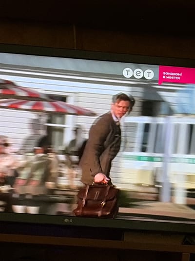 The image is a screenshot of a television screen showing a man walking with a briefcase. He is wearing a dark suit jacket, a light-colored shirt, and a tie. The man is in motion, with his body angled to the side and his head turned slightly towards the camera. He is carrying a dark brown leather briefcase in his right hand.
In the background, there is a building with horizontal siding, possibly a house or a storefront. There are also elements of what appears to be a patio or outdoor seating area, including a red and white striped umbrella and some tables and chairs that are blurred due to motion.
In the top right corner of the screen, there is a pink banner with white text that reads "закохані в життя" which translates to "in love with life." To the left of this banner, there is a logo with three white circles containing the letters "т е т". In the bottom left corner of the television screen, there is an LG logo.
The overall scene suggests a casual outdoor setting, possibly in a town or city. The presence of the television screen and the on-screen graphics indicate this is a broadcast program. The image quality is somewhat grainy, as expected from a photograph of a television screen.
The mention of Osolynka, Ukraine, in the prompt is not verifiable from the visual information provided in the image.