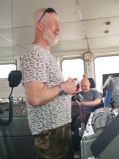 An indoor shot captures two men in what appears to be the bridge or wheelhouse of a ship, set in Городище, Russia.

In the foreground, a fair-skinned man with light gray hair and a prominent beard stands smiling, looking towards the right. He wears sunglasses on his head, a light-colored short-sleeved t-shirt patterned with grey fish, dark patterned trousers, a metal chain, and a wristwatch. He holds a glass cup containing a dark liquid, likely tea, with a spoon or stirrer. To his left, a black "Mercury" brand microphone with a coiled cord is mounted on a control console featuring various buttons and dials.

In the mid-ground, a bald man with fair skin is seated in a black chair, wearing a striped short-sleeved t-shirt. He looks down, engrossed in a small device, possibly a mobile phone. Behind him, parts of the ship's controls, including large red, blue, and gray levers, are visible.

The setting features large windows in the background, revealing an overcast sky, suggesting daytime. A round wall clock mounted above the windows shows the time to be approximately 11:30. The ceiling is white and paneled with metal strips and hooks. On the left, papers with visible but illegible Cyrillic text and a diagram are attached to a board.

The scene depicts a casual moment, possibly a break, within the utilitarian environment of a vessel's command deck. The standing man appears relaxed and content, while the seated man is focused on his device.