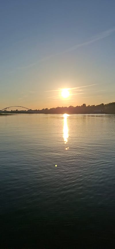 The image captures a serene sunset over the Pärnu River in Pärnu, Estonia. The sun, a bright orb, is positioned slightly left of center in the upper portion of the frame, casting a shimmering reflection that stretches across the water's surface. The river occupies the majority of the image, its gentle ripples catching the golden light. On the far bank, a tree line forms a dark silhouette against the horizon. To the left, a distinctive arched bridge spans the river, its structure faintly visible against the fading daylight. The sky above is a gradient of pale blue transitioning to a soft orange near the horizon, with faint wisps of clouds. The overall scene is one of tranquility and natural beauty.