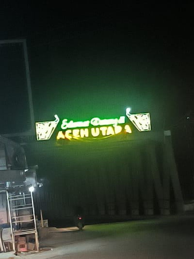 At night, the image depicts a roadside establishment in Kotapantonlabu, Indonesia, displaying a neon sign that reads "Selamat Datang di ACEH UTARA." The sign is illuminated in green and yellow, featuring stylized images of iced tea glasses on either side of the text. A simple metal structure, possibly a small kiosk or food stall, is visible on the left, with some visible contents. The surrounding environment suggests an urban or semi-urban setting.