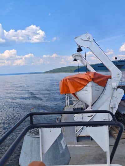 The image depicts a view from the deck of a boat on a wide river under a partly cloudy blue sky. In the foreground, a white lifeboat, covered by an orange tarp, is secured by a davit crane system. The crane is white and appears to be made of metal. Part of the boat's railing, a black metal bar, is visible in the lower left quadrant of the image.
The river is calm with gentle ripples reflecting the sky. In the distance, a forested, hilly shoreline stretches across the horizon. A small vessel can be seen in the middle ground on the river. The overall scene suggests a serene day on the water, likely during daytime.
While the image does not explicitly show the city of Podkamennaya Tunguska, Russia, the described elements of a wide river, forested shores, and riverine transport are consistent with the geographical characteristics of the region.