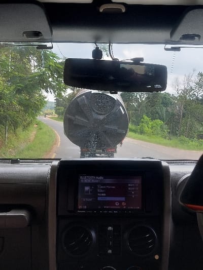 The image depicts a scene from inside a vehicle looking forward through the windshield on a road in Kisanga, Tanzania. The interior visible includes the overhead console with two sun visors, a rearview mirror, and a dashcam mounted below the mirror. The dashboard and center console are prominent, featuring an infotainment screen displaying "BLUETOOTH Audio," "Diamond Platinumz" as the artist, "Sound" settings, and "iPhone" as the connected device, with the time "21:28" visible.
Outside the vehicle, a paved road with green vegetation lining both sides is visible under an overcast sky. Directly ahead, a large, dark-colored, horizontally ribbed cylindrical tank, consistent with a water or liquid storage tank, is being transported on the rear of a flatbed truck or trailer. The rear surface of the tank faces the camera. In the distance, on the left side of the curving road, a small, indistinct figure, possibly a person on a motorcycle or bicycle, is visible.