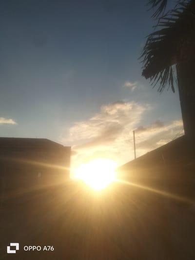 The image depicts a bright sunset with the sun positioned low on the horizon, partially obscured by buildings and a palm tree. The sky is a gradient of blue and orange, with scattered clouds reflecting the sunlight. In the foreground, a dark building with a sloped roof is visible on the left, and a fence runs along the right side of the frame. A palm tree's fronds are silhouetted against the sky on the upper right. The sun's rays create a lens flare effect, illuminating the scene with a warm glow. The OPPO A76 logo is present in the lower left corner. The image appears to be taken outdoors, likely in a residential or urban area with vegetation. The specific location in Babatan, Indonesia, is not visually identifiable from the image itself.
