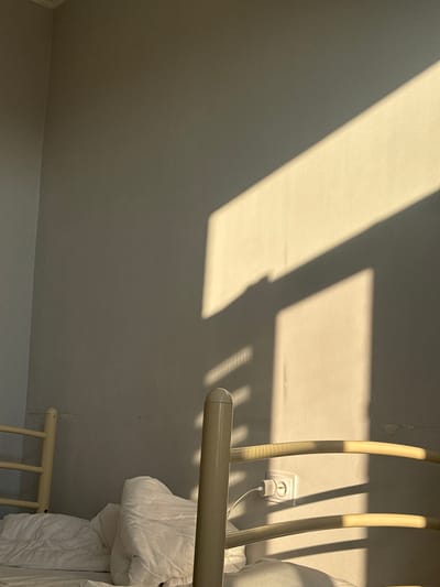 The image depicts a close-up view of a bed in a room, with sunlight casting strong shadows across a pale wall. In the foreground, a portion of a light-colored bed frame is visible, along with white bedding. A white electrical outlet with a connected charger is mounted on the wall to the right of the bed frame. The wall itself appears to be painted a neutral color and shows some minor imperfections. The lighting suggests it is either early morning or late afternoon, with the sun's rays entering from the side. The location of Chemerivtsi, Ukraine is mentioned, but there are no specific visual cues within the image to confirm this location. There are no people visible in the image, and no actions or interactions are taking place.
